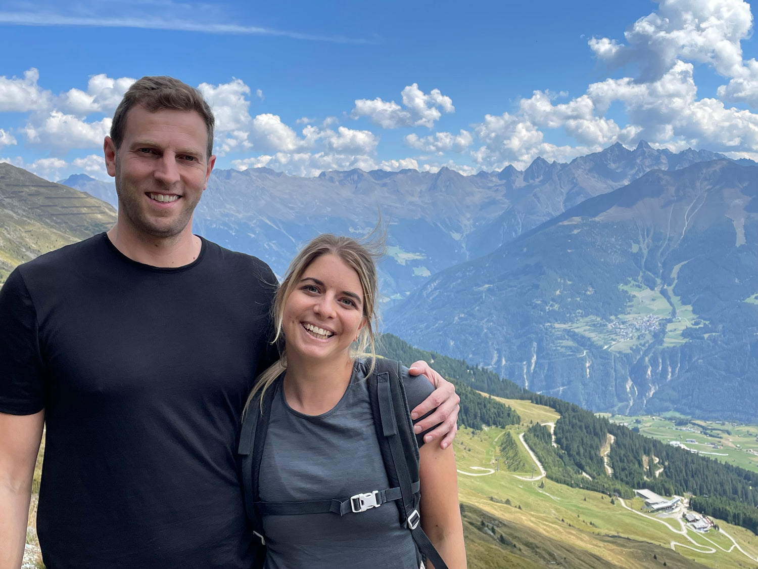 Ein lächelnder Mann und eine lächelnde Frau stehen auf einem Bergpfad mit Blick auf ein malerisches Tal. Die Landschaft besteht aus sanften Hügeln, gewundenen Straßen und fernen Bergen unter einem teilweise bewölkten Himmel. Beide sind leger gekleidet und wirken glücklich.