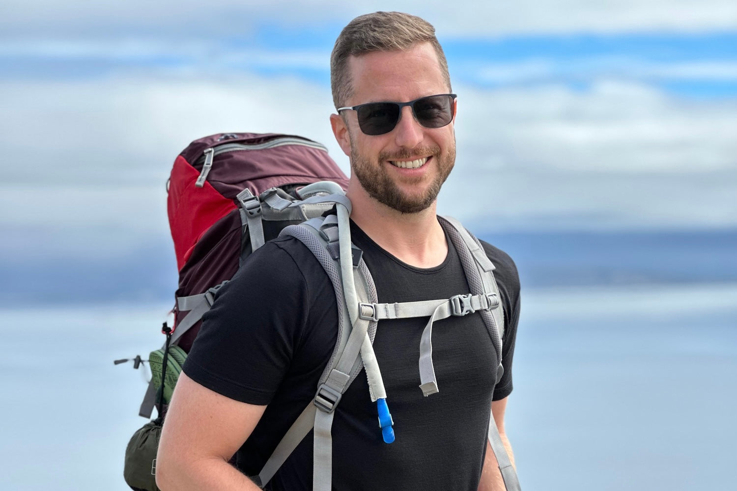 Eine Person mit Sonnenbrille und schwarzem T-Shirt lächelt und trägt einen großen Rucksack mit rotem Überzug. Im Hintergrund sind ein bewölkter Himmel und eine verschwommene Landschaft zu sehen.