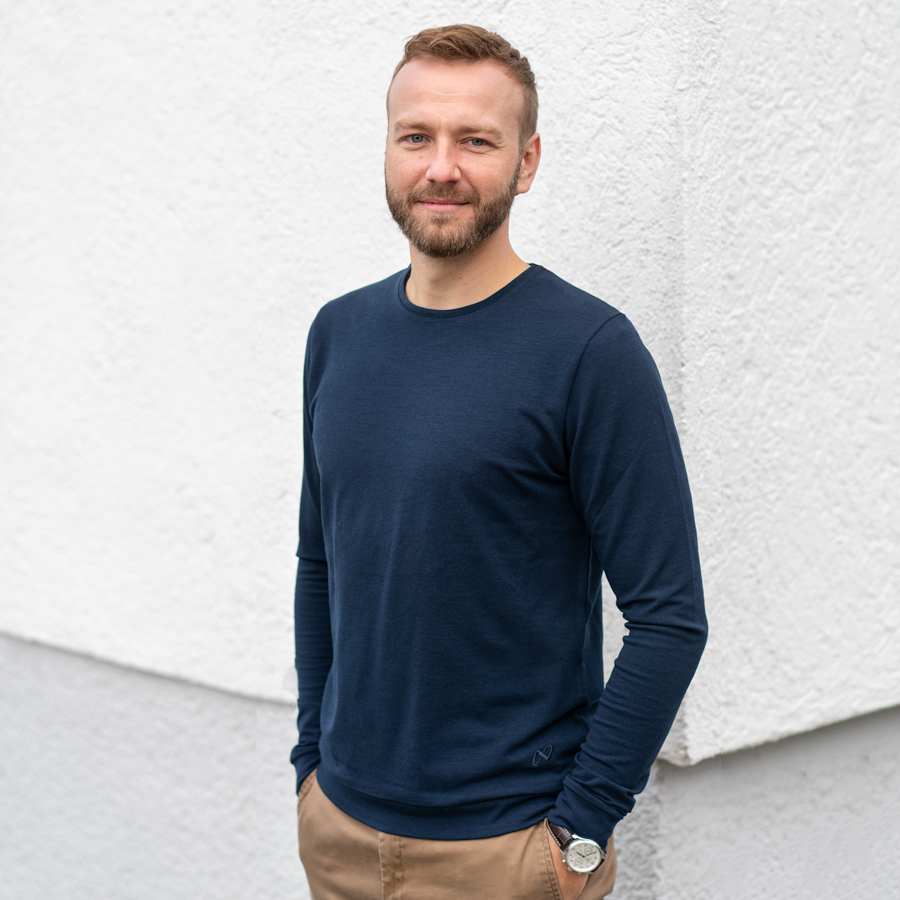 Ein Mann mit Bart, der einen marineblauen Pullover und hellbraune Hosen trägt, steht vor einer weißen strukturierten Wand. Er hat die Hände in den Taschen und lächelt in die Kamera.