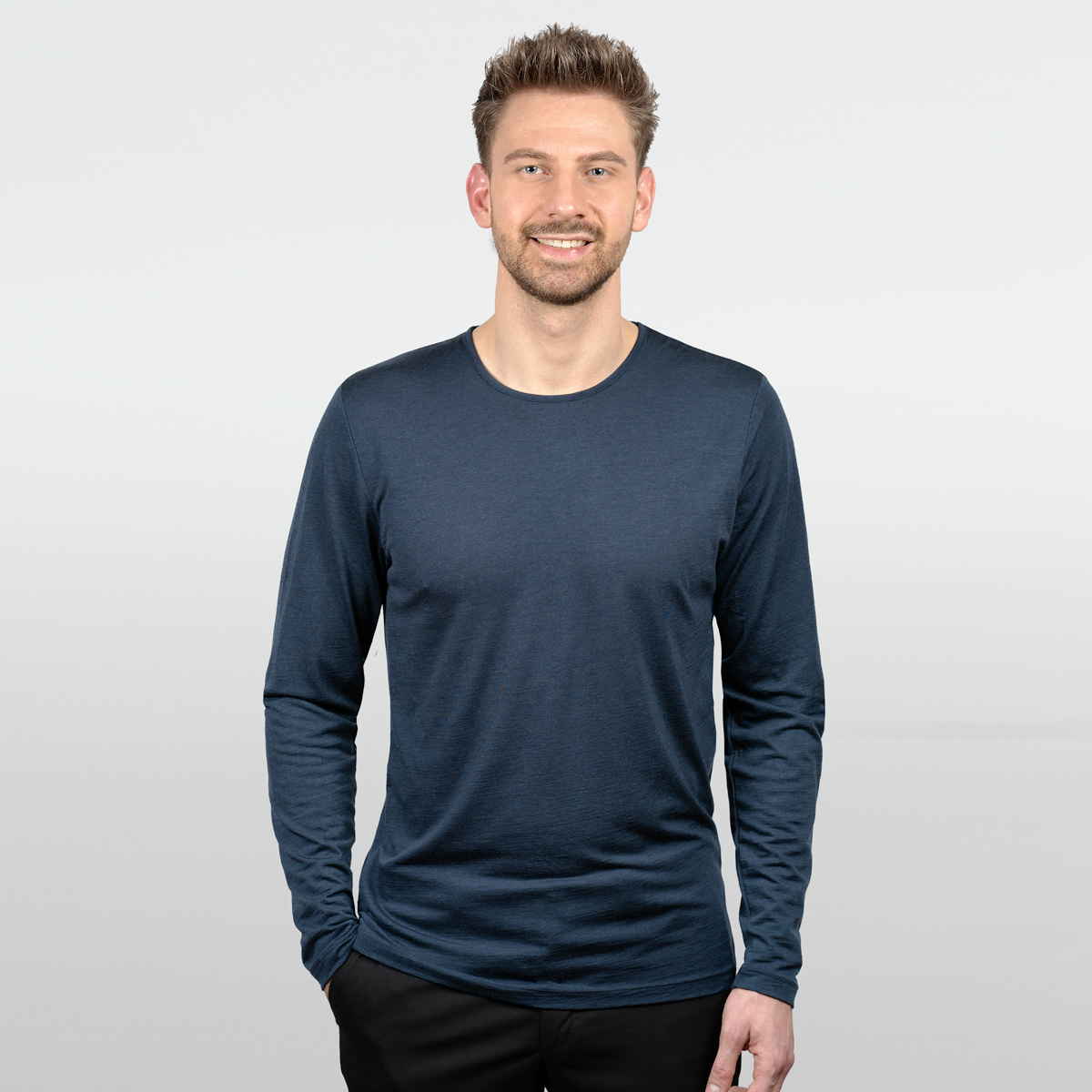 Eine Person mit kurzen Haaren lächelt in die Kamera. Sie trägt ein langärmeliges, dunkelblaues Hemd und eine schwarze Hose vor einem schlichten weißen Hintergrund.