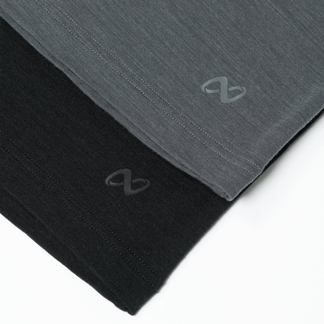 Zwei gefaltete T-Shirts in Grau und Schwarz liegen nebeneinander auf weißem Hintergrund. Jedes Shirt hat ein dezentes, geschwungenes Logo am Saum aufgedruckt.