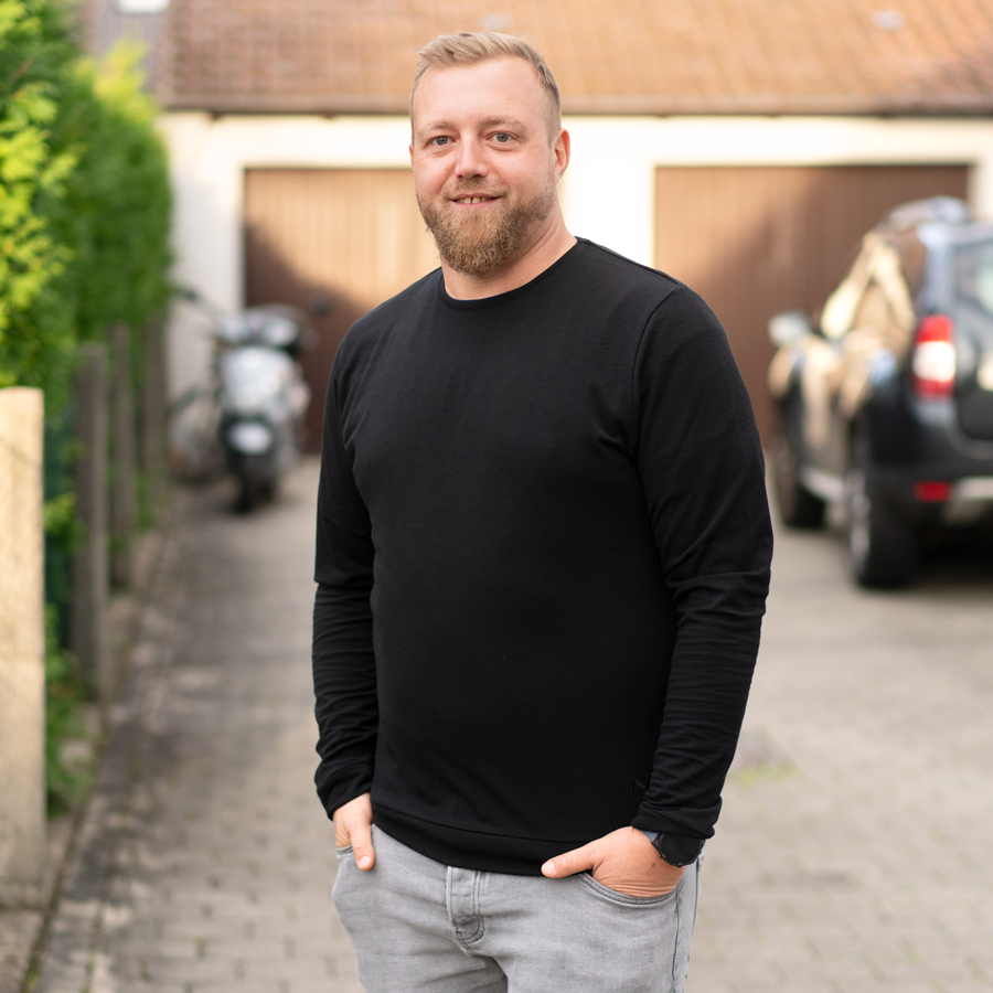 Ein Mann mit Bart steht draußen auf einer gepflasterten Auffahrt und trägt ein schwarzes Langarmhemd und graue Hosen. Die Hände in den Taschen lächelt er in die Kamera. Im Hintergrund sind Autos und Grünflächen unter einem bewölkten Himmel zu sehen.